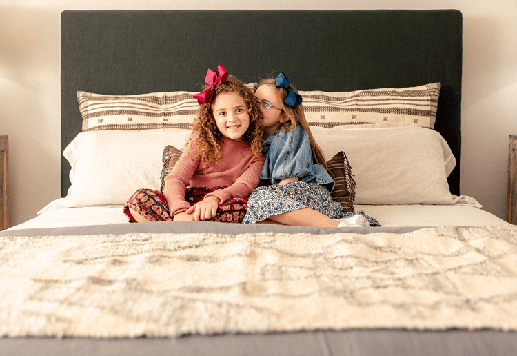 Photo of two sisters whispering secrets to each other while sitting on a bed in their Betenbough home.
